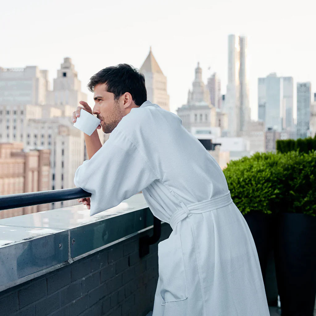 A man in a white bathrobe stands on a balcony, drinking from a white mug, with a city skyline in the background.