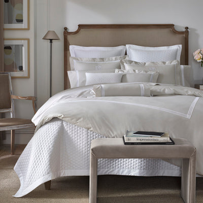 Sferra Ritz ivory and white cotton bedding set on upholstered bed with wooden headboard.