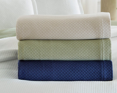 Three folded textured blankets in beige, green, and navy are stacked on a bed with a white quilted cover.