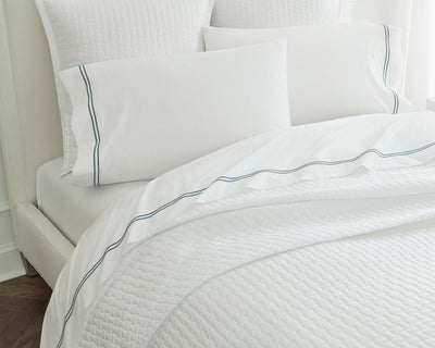 A neatly made bed with white bedding, a quilted coverlet, and pillowcases featuring a simple blue stripe along the edge.