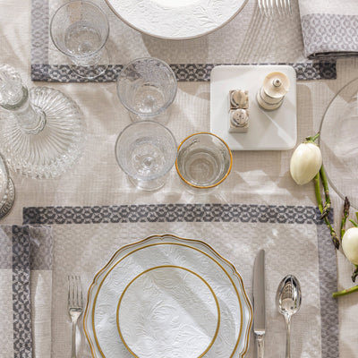 Évasion Céleste ivory linen tablecloth with grey border, set with crystal and gold-rimmed china.