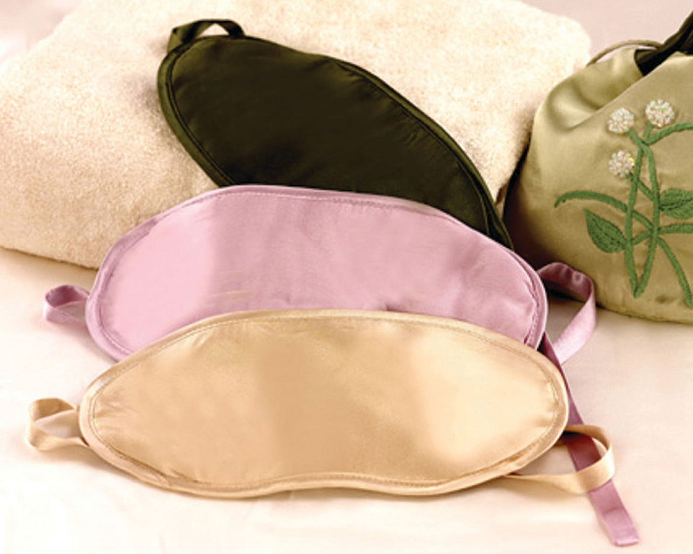 Three satin sleep masks in black, light purple, and beige are displayed on a towel with a green embroidered bag in the background.