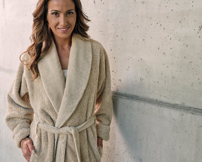 A woman stands against a concrete wall, wearing a beige belted robe with her hands in the pockets, and smiles at the camera.