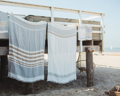 Striped towels hang from a wooden deck above a sandy beach, with a basket nearby and a sailboat visible in the distance on the water.