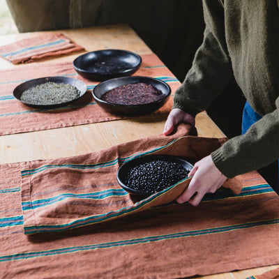 Rust linen table runner with teal stripe, styled on wood table with bowls of grains.