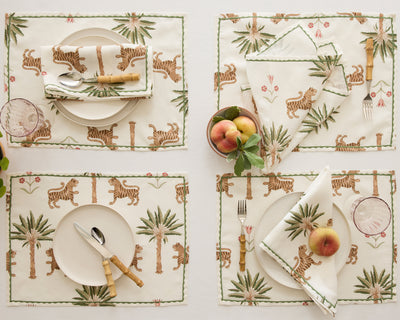 A table set with four place settings featuring plates, cutlery, and napkins with a tiger and palm tree pattern. A bowl of peaches and individual peaches decorate the table.