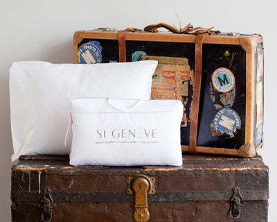 Two stacked vintage suitcases with travel stickers, a white pillow, and a white bag labeled St GENEVE placed in front of them.