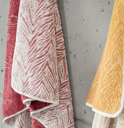 Vasco crimson and saffron patterned terry bath towels hanging against concrete wall.