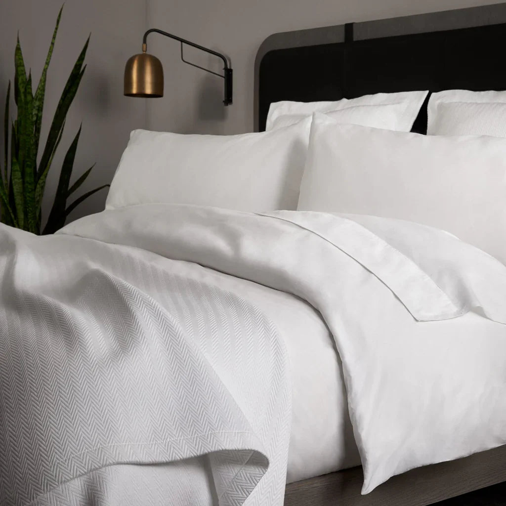 A neatly made bed with white pillows, a white comforter, and a textured white blanket, next to a potted plant and a brass wall-mounted lamp.