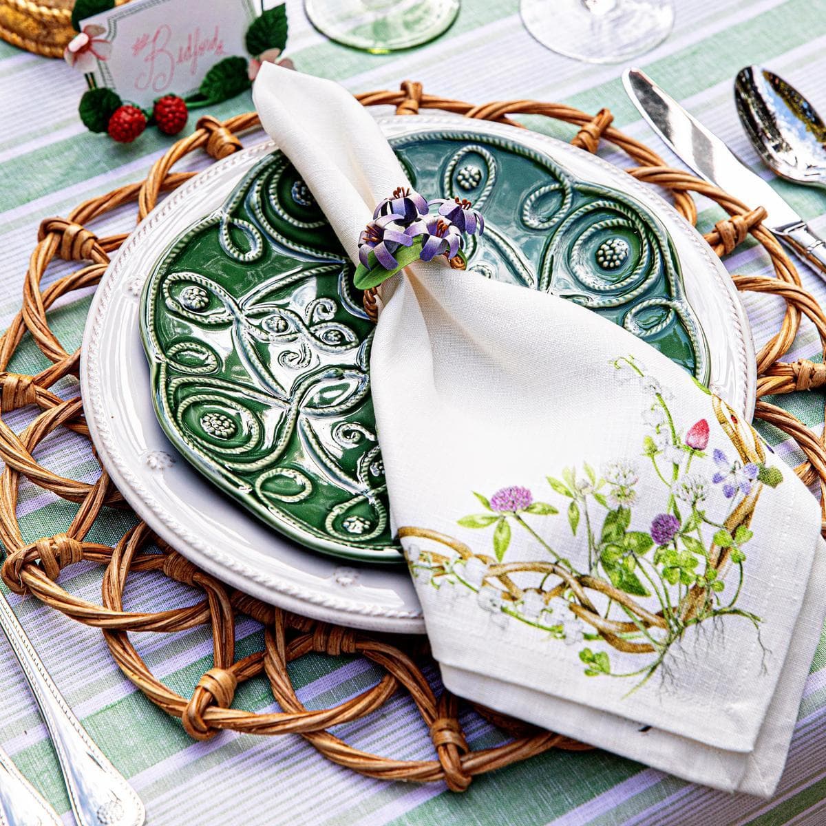 White linen napkin with embroidered wildflower meadow motif, styled open beside a green ceramic plate.