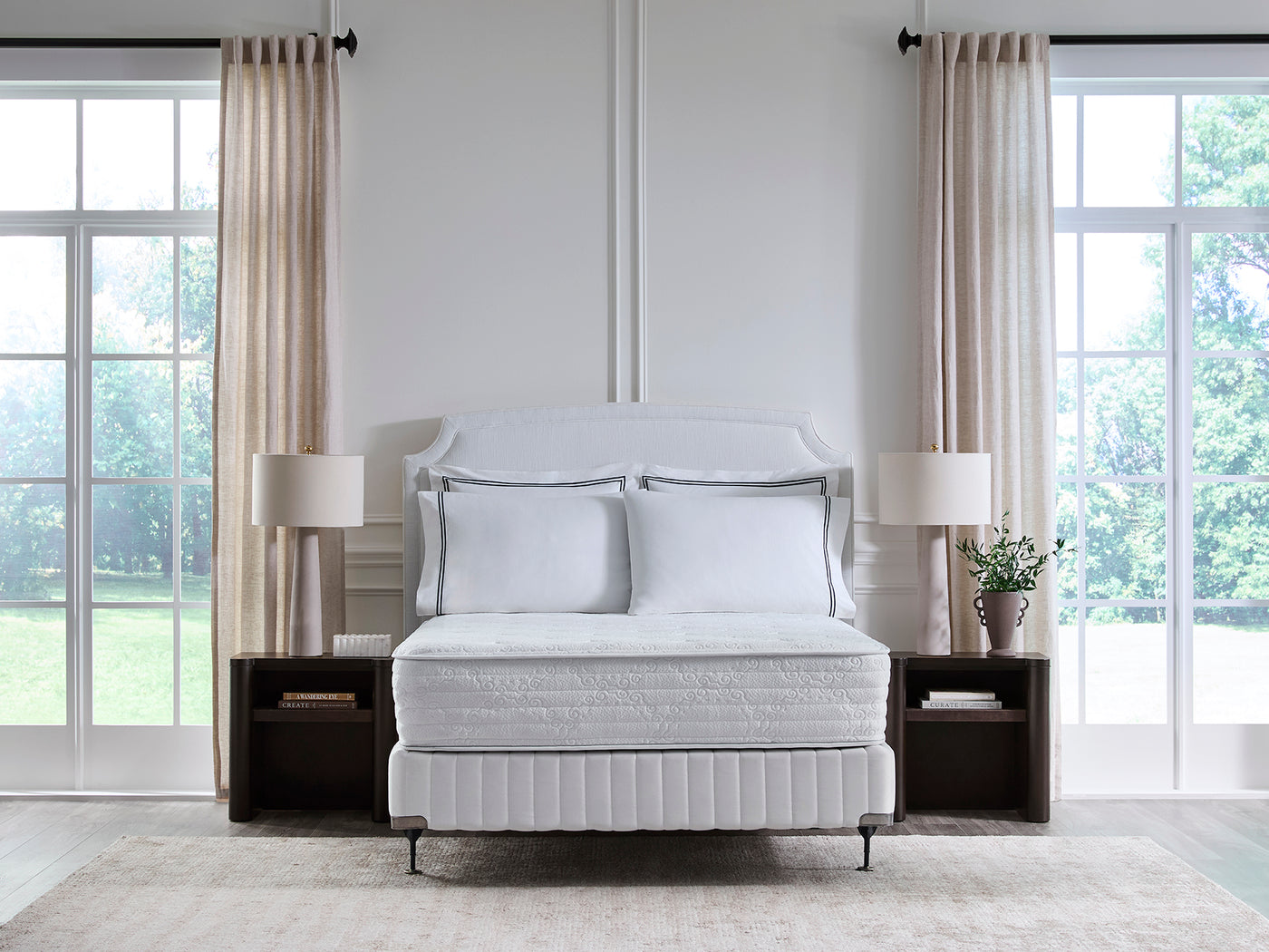 A white upholstered bed with matching pillows and a quilted mattress sits centered in a bright, modern bedroom with tall windows, beige curtains, and dark wood nightstands.