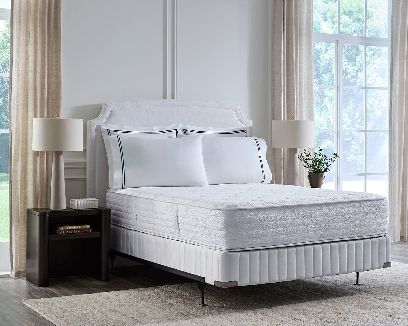 A white upholstered bed with a quilted mattress and matching pillows in a bright, modern bedroom with beige curtains, bedside tables, lamps, and a light rug.