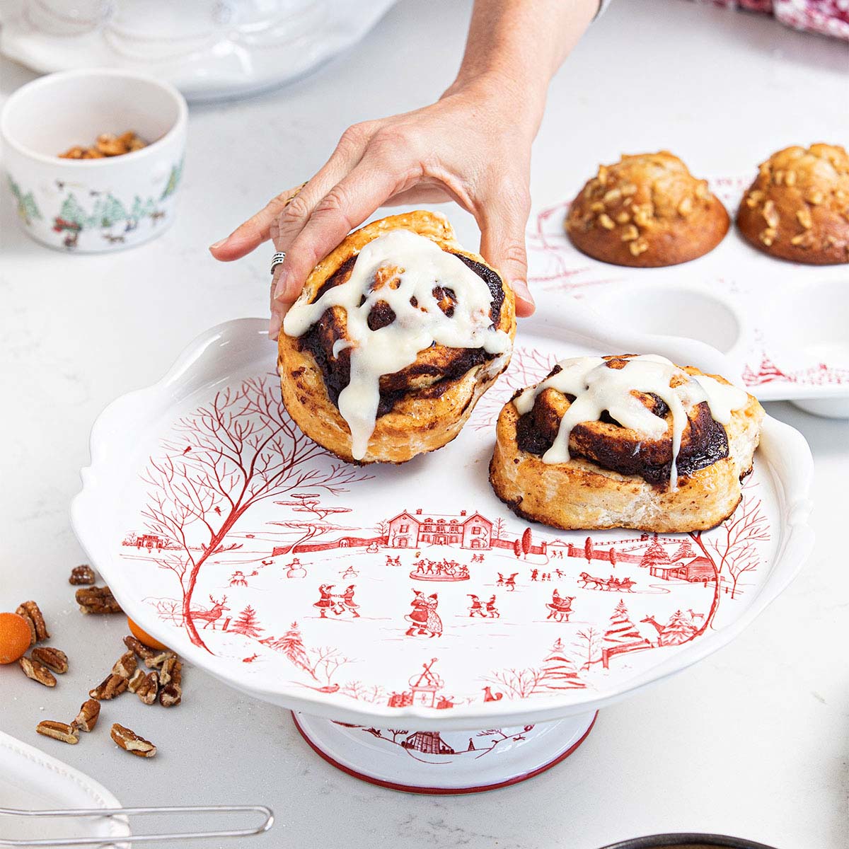 Hand placing cinnamon buns on a ruby Winter Frolic cake stand