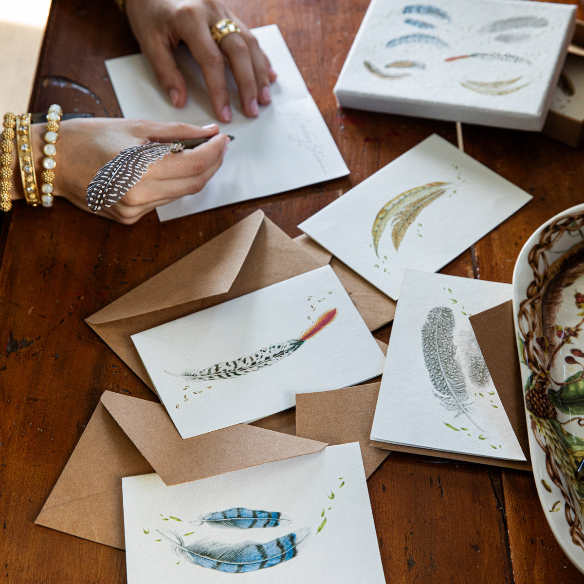 Hands writing Forest Walk feather note cards on a wooden table with kraft envelopes