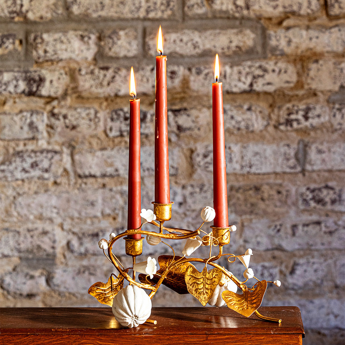 Forest Walk gold pumpkin and vine triple candlestick holder with red tapers on a table against a brick wall