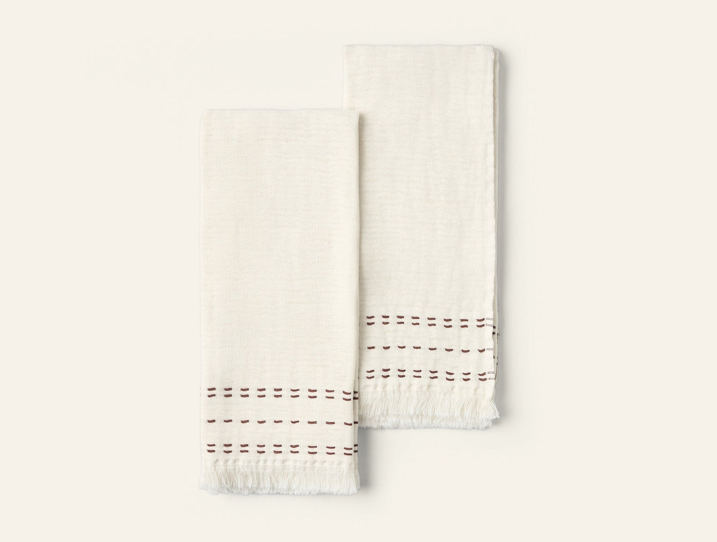 Pair of ivory hand towels with fringed hems and burgundy embroidered stitch border detail.