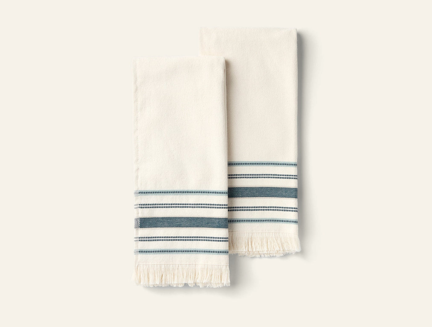 Close-up of two ivory Coyuchi Aptos hand towels with teal stripes and fringe hem.