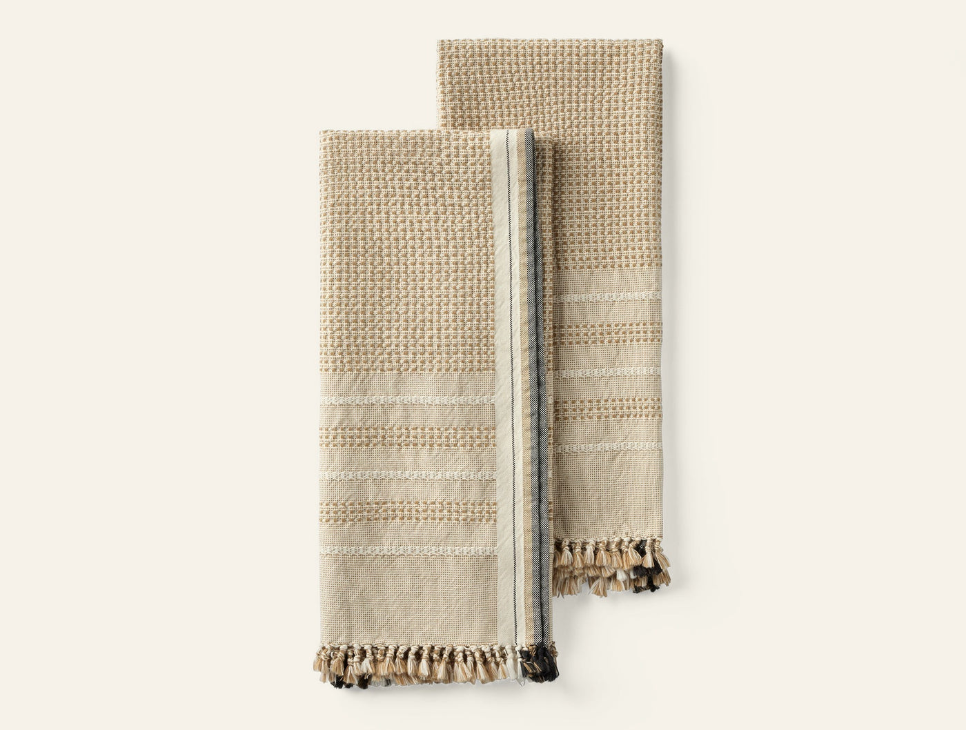 Two folded Coyuchi Bay hand towels in natural with waffle texture, stripe detail, and fringe hem.