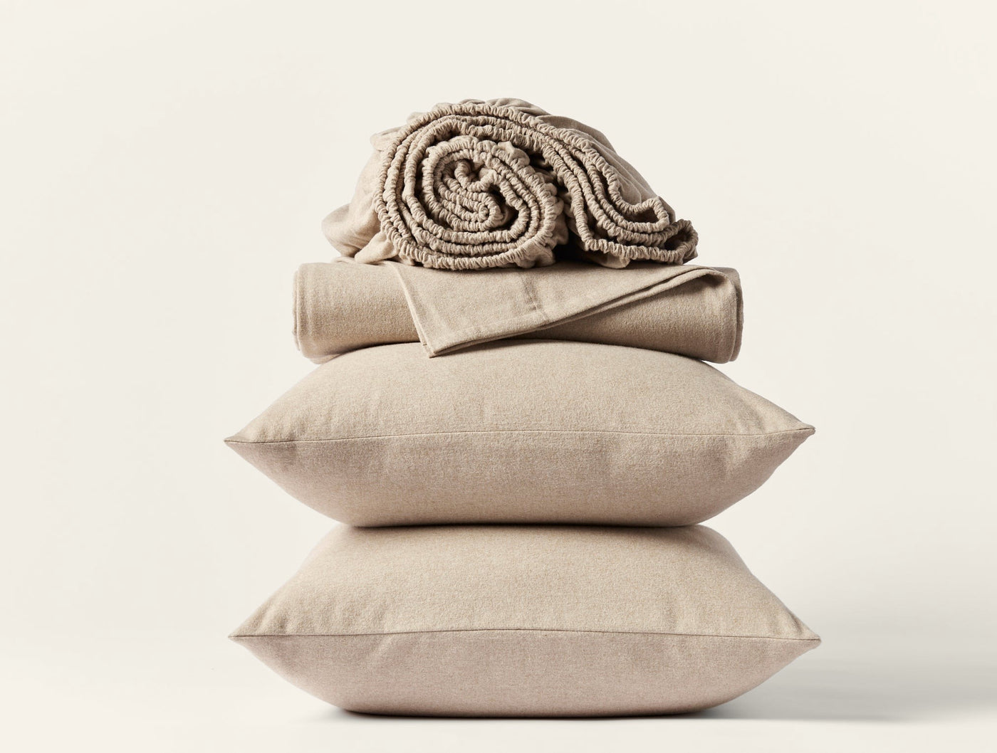 Stack of two sand-colored linen pillows topped with folded flannel sheets and a rolled textured blanket.