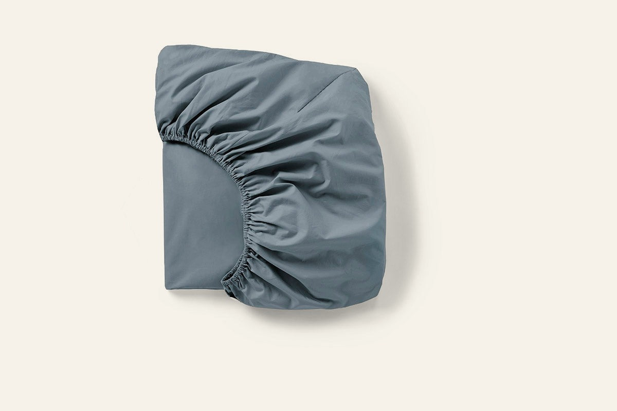 Coyuchi Cloud Soft Sateen fitted sheet folded, showing elasticated corners in slate blue.