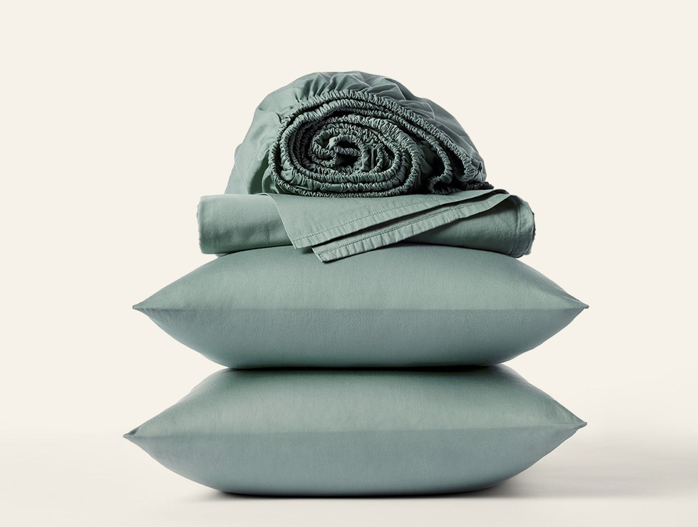 Sage green sateen sheet set styled with stacked pillowcases and rolled fitted sheet.