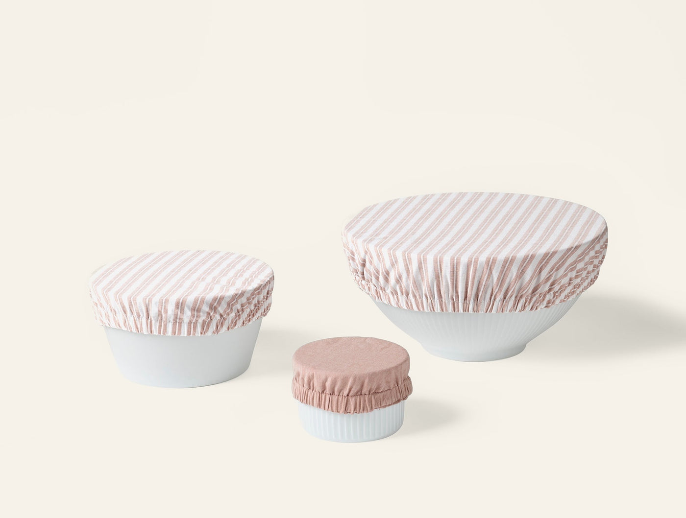 Three Coyuchi bowl covers in blush stripe and solid blush, covering white bowls in small, medium, large sizes.