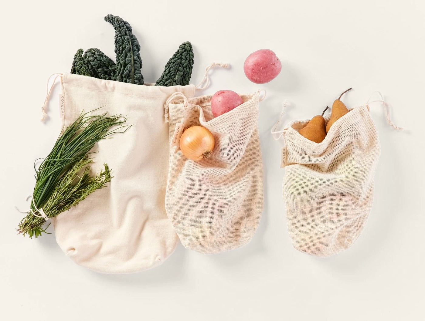 Three natural cotton Coyuchi produce bags in varied sizes, filled with fresh vegetables and fruit.