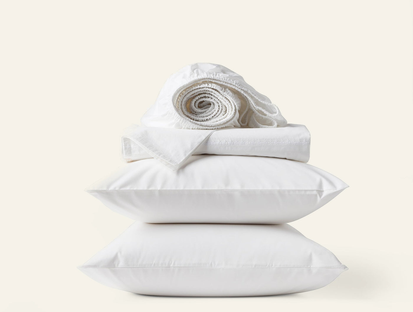 Coyuchi Floret Embroidered sheet set in white, stacked with pillows and rolled sheets.