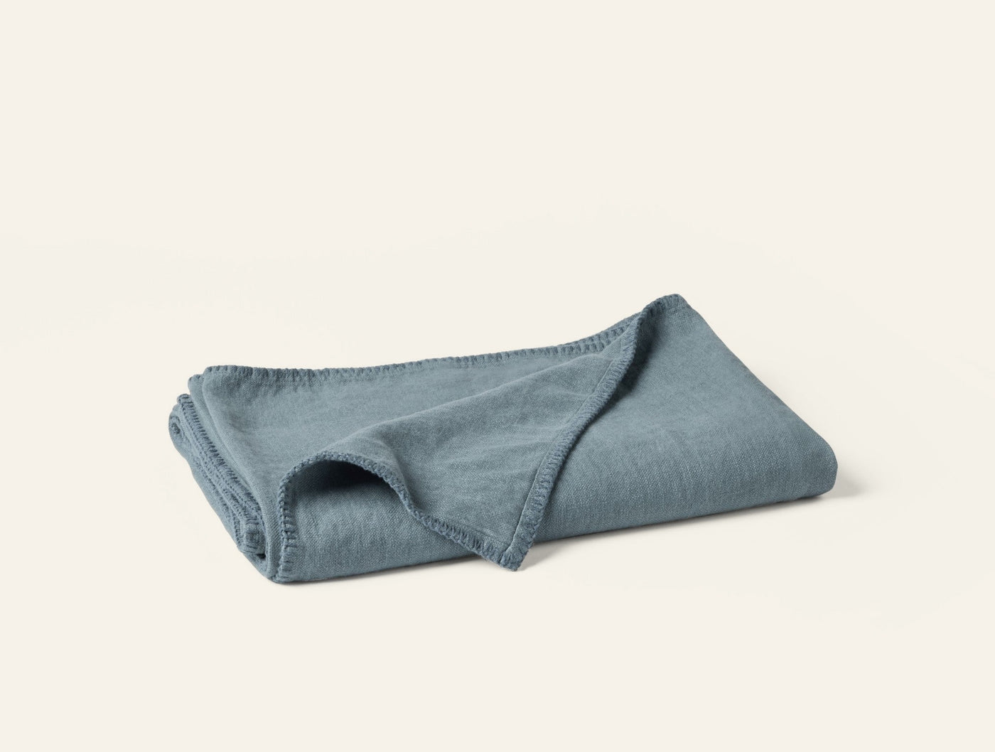 Folded slate blue linen throw, corner turned back to show texture and blanket-stitched edge.