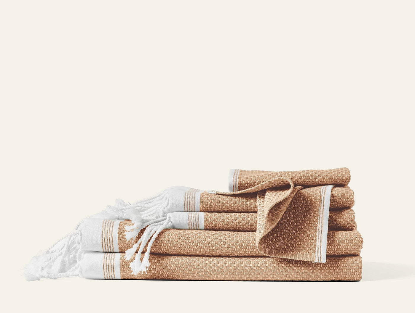 Stacked camel and white waffle-weave towel set with fringed hammam towels on left.