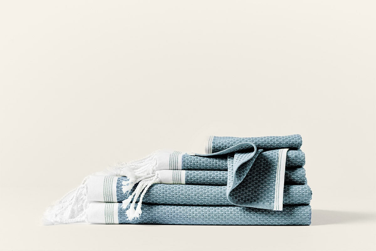 Stacked slate blue Coyuchi Mediterranean towels with white fringe and woven texture detail.