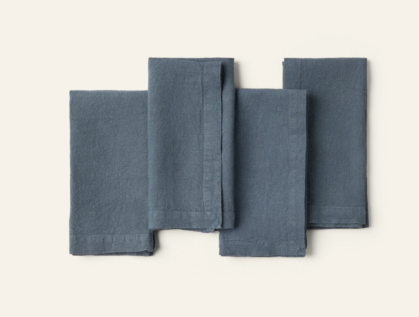 Four slate blue linen napkins arranged in a flat lay, slightly overlapping on a white background.