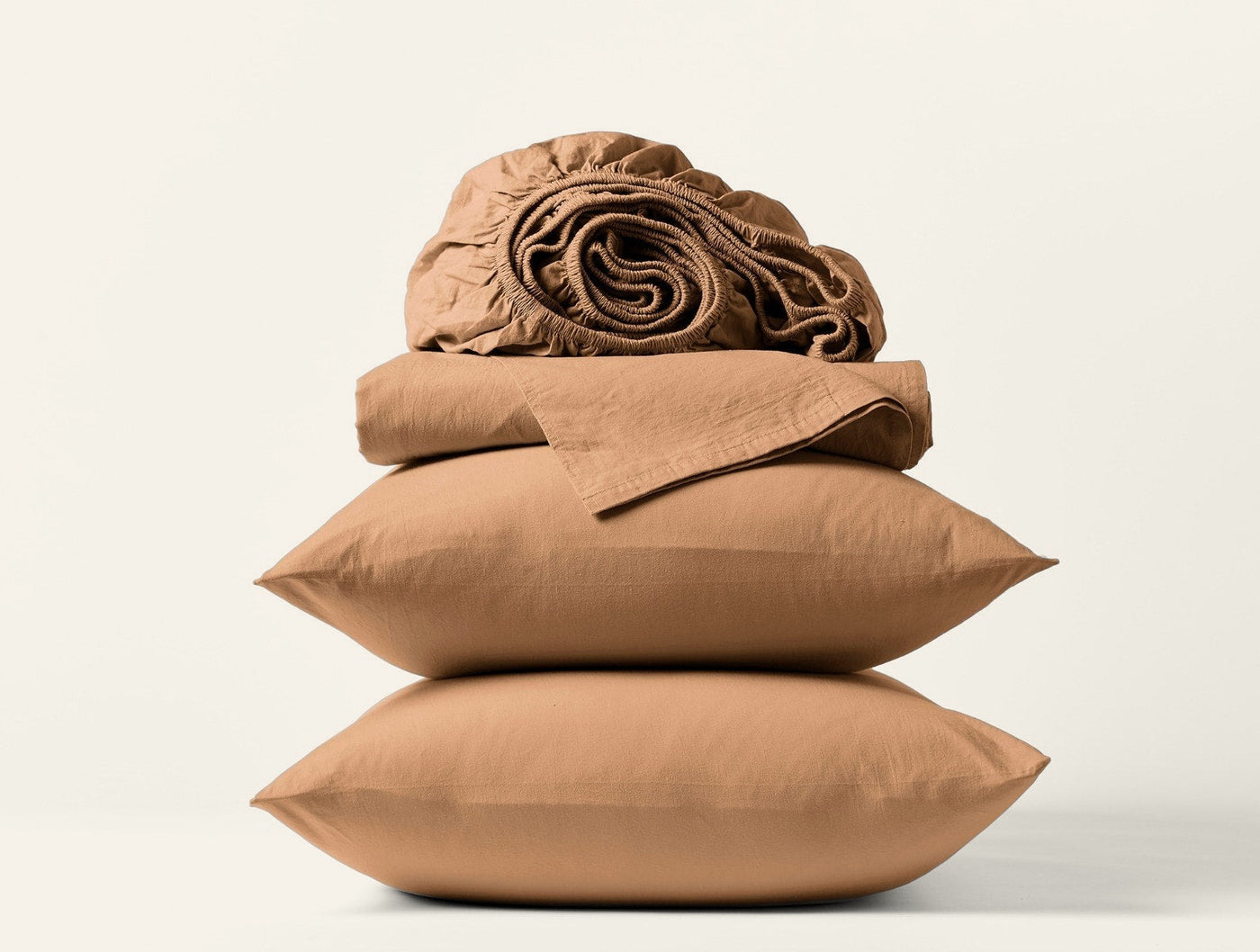 Close-up of stacked caramel Coyuchi Crinkled Percale pillowcases and folded sheets on white background.