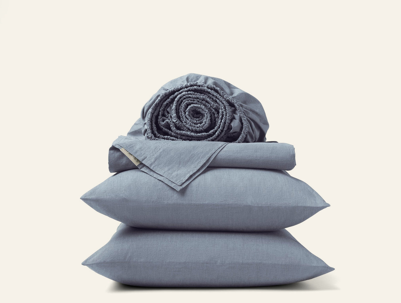 Stacked slate blue Coyuchi Crinkled Percale pillowcases and sheets with a rolled fitted sheet on top.