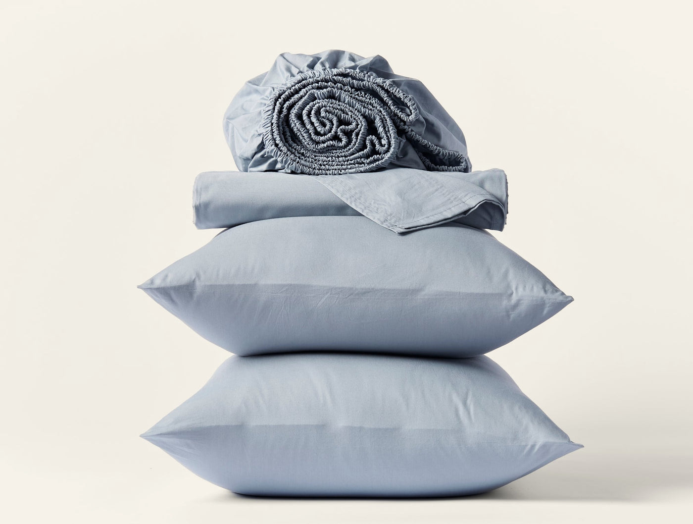 Stacked slate blue Coyuchi percale pillowcases and folded fitted sheet on white background.