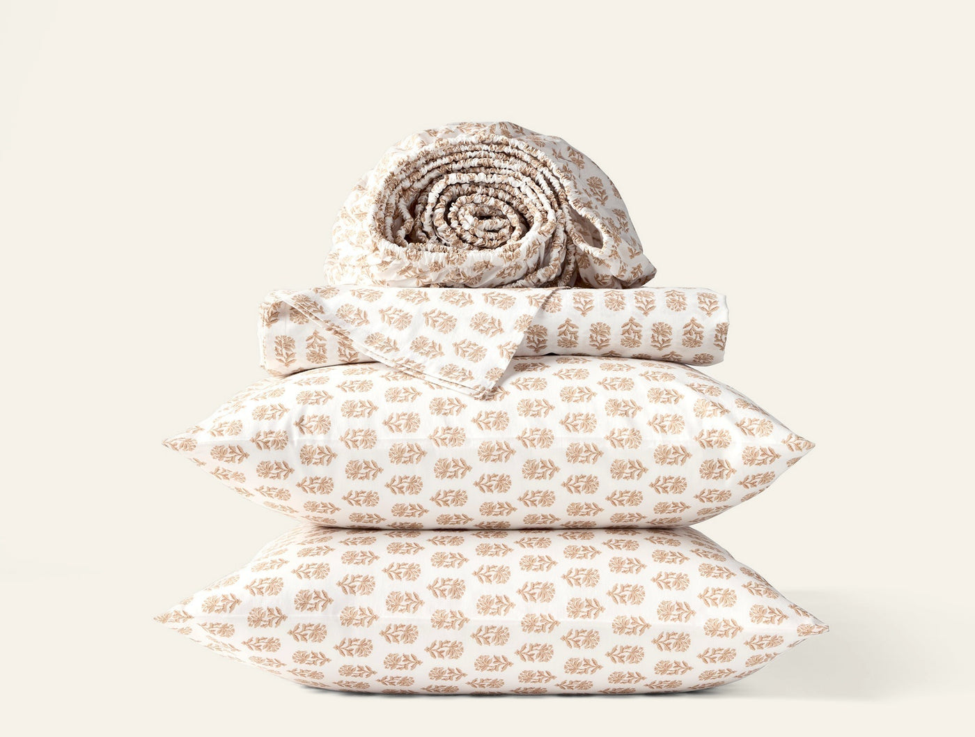 Stacked Coyuchi pillowcases in warm sand block-print pattern, with rolled sheet set on top.