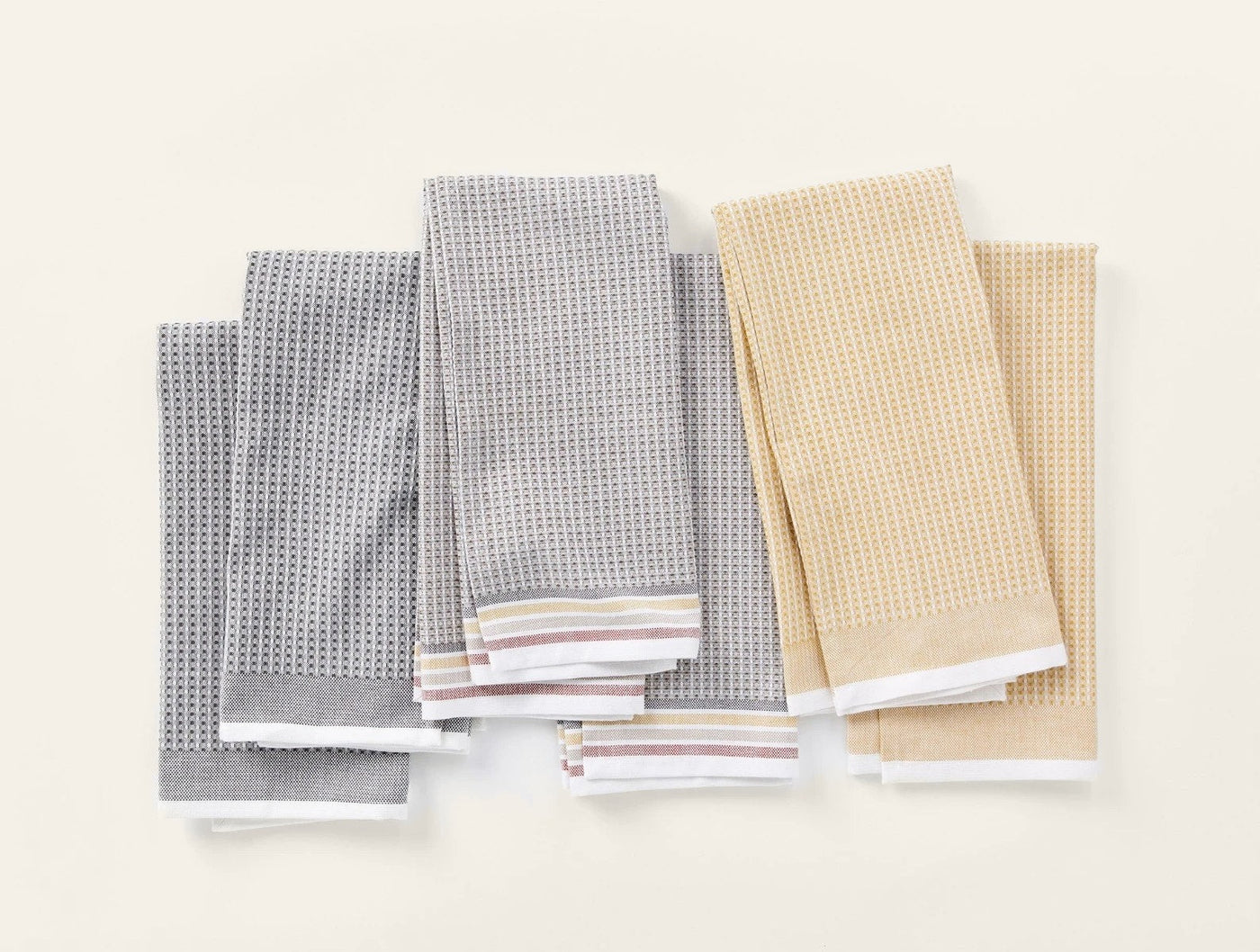 Six Coyuchi Waffle kitchen towels folded flat, three grey and three gold, with striped hems.