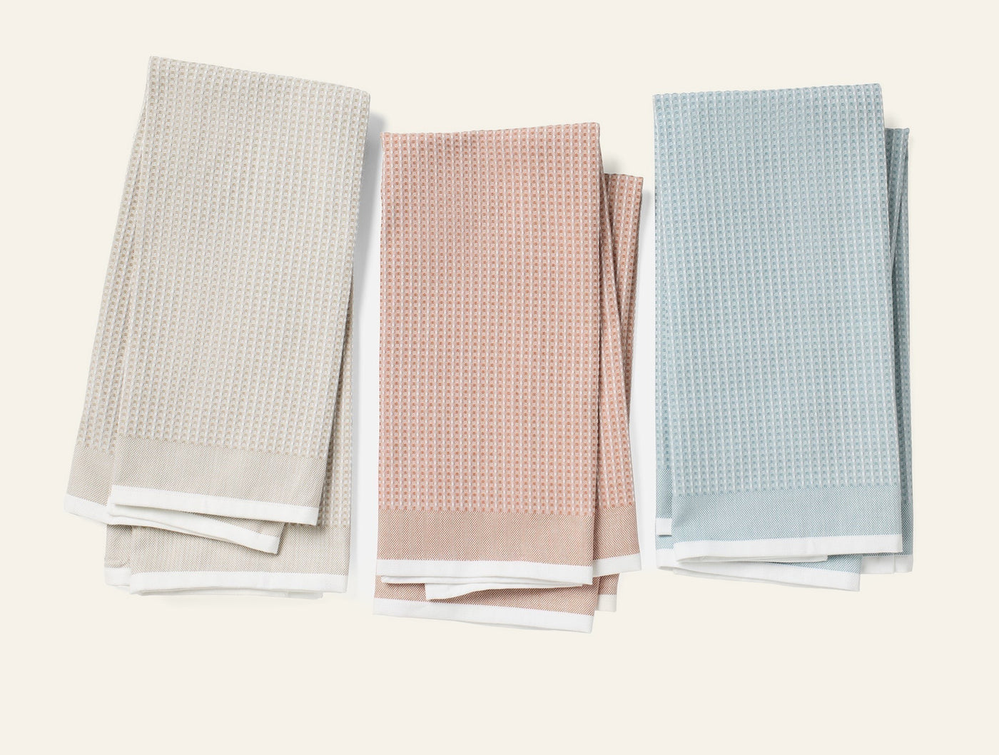Three folded Coyuchi waffle-weave kitchen towels in stone, blush, and sky blue with white borders.
