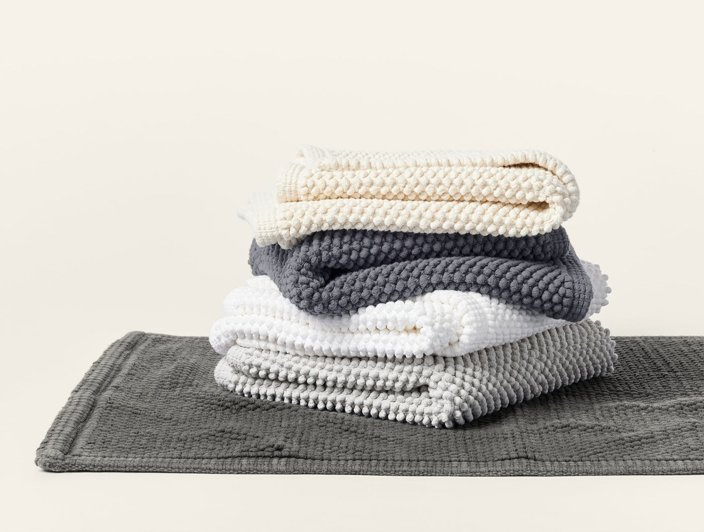 Coyuchi Pebbled bath rugs stacked in ivory, charcoal, white, and slate colorways.