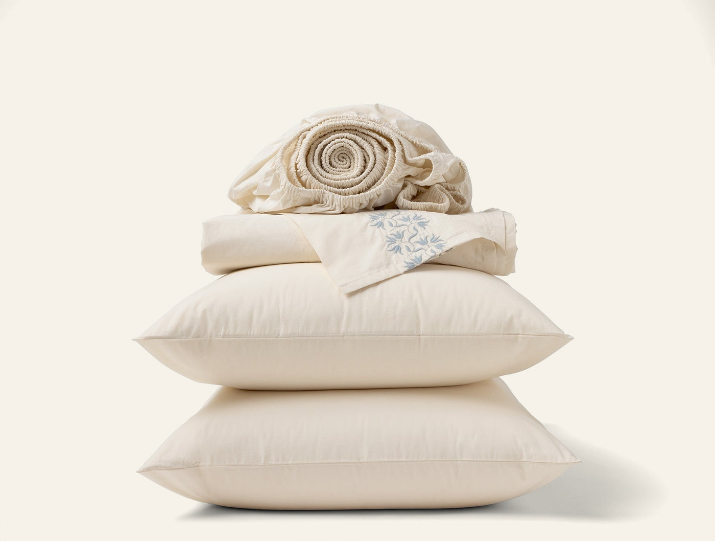 Close-up of stacked ivory Coyuchi Refined Percale pillows and folded sheets on white background.