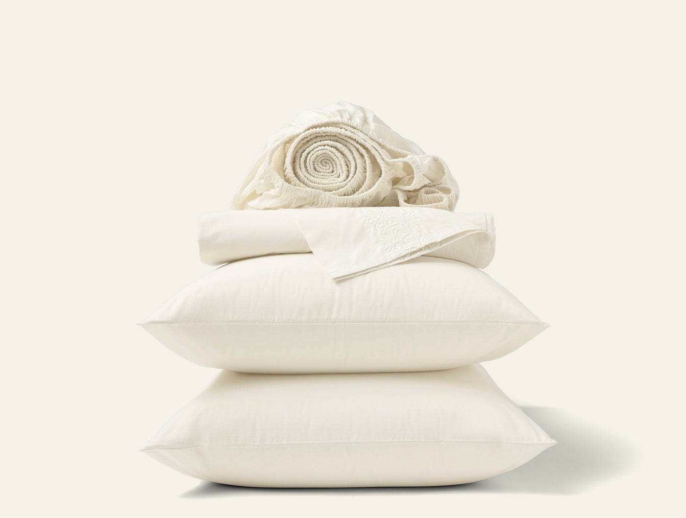 Coyuchi Refined Percale sheet set styled as stacked ivory pillows with rolled flat sheet on top.