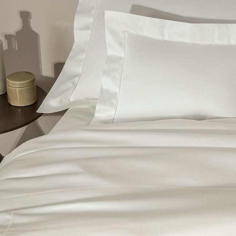 A neatly made bed with Frette's Single Ajour | Sheet Set in white cotton sateen, alongside a round bedside table holding a gold container and a glass bottle.