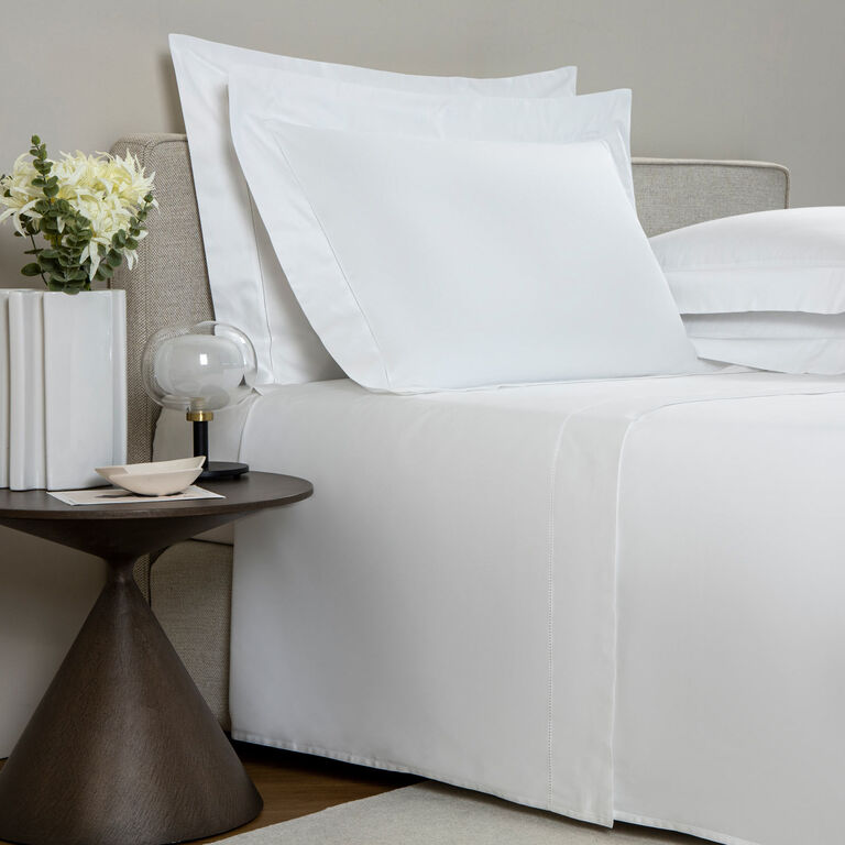 A neatly made bed with Frette's Single Ajour Sheet Set in white cotton sateen and open hemstitch sits beside a round side table topped with a vase of flowers, books, a lamp, and a small dish.