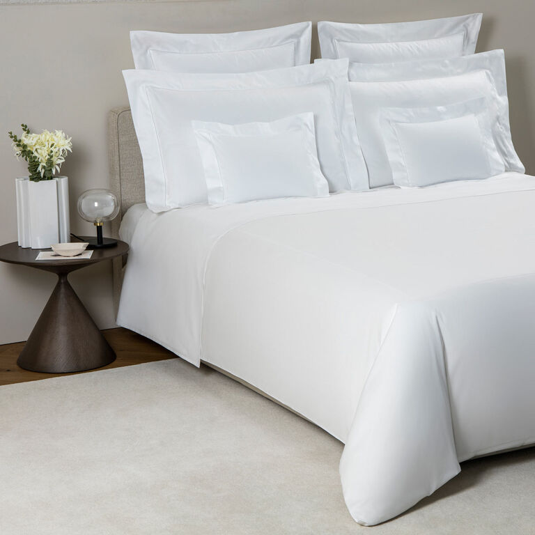 A neatly made bed with Frette’s Single Ajour Pillowcase Set in white cotton sateen, featuring open hemstitch details, sits beside a round side table with a lamp, white flowers, and a book on a light-colored rug. Made in Italy.