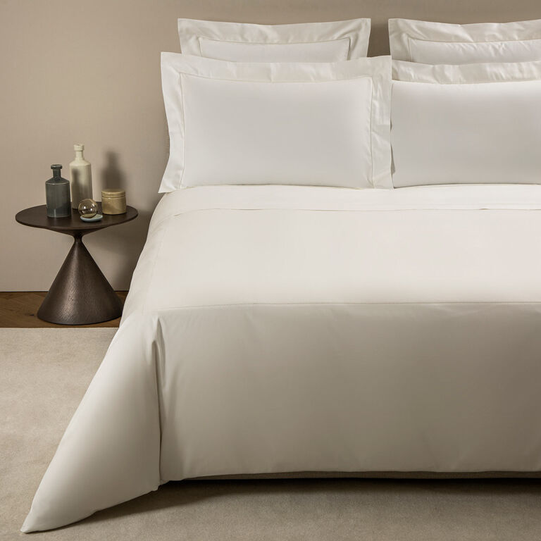 A bed dressed in Frette’s Single Ajour Pillowcase Set, featuring white cotton sateen with open hemstitch detail, is complemented by four pillows and sits next to a round table with decorative bottles and a glass.