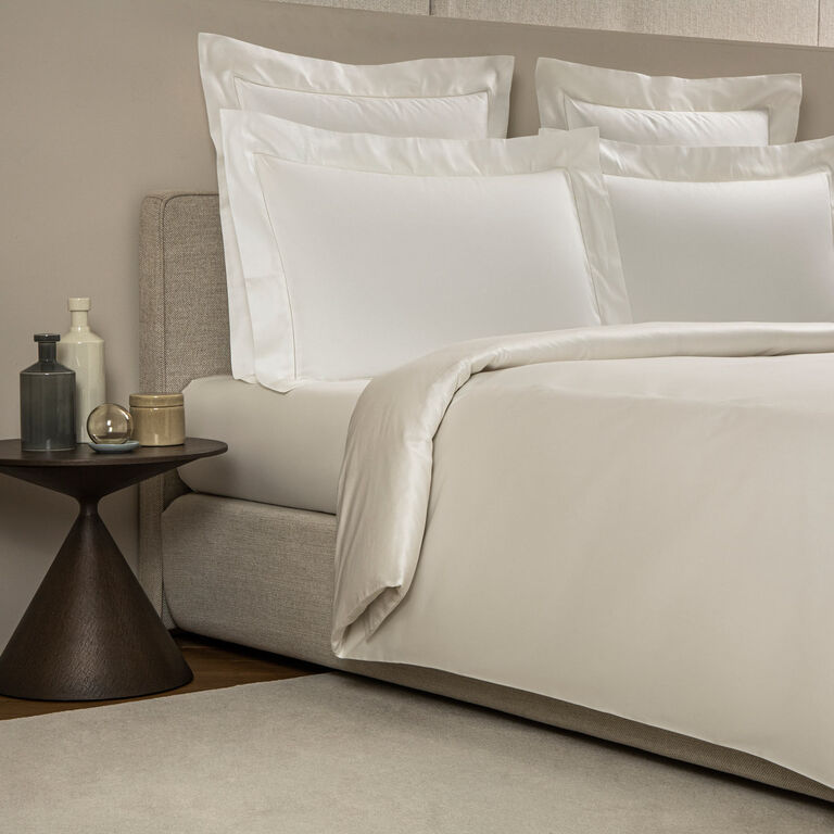 A bed is styled with Frette's Single Ajour Pillowcase Set featuring white cotton sateen and open hemstitch details, beside a round wood side table topped with decorative vases and a cup.