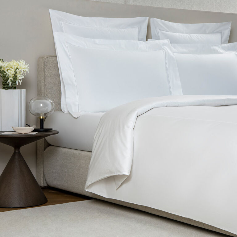 A neatly made bed features Frette’s Single Ajour Pillowcase Set in white cotton sateen with an open hemstitch, beside a small round table with a vase of white flowers, a lamp, and a cup. Made in Italy.