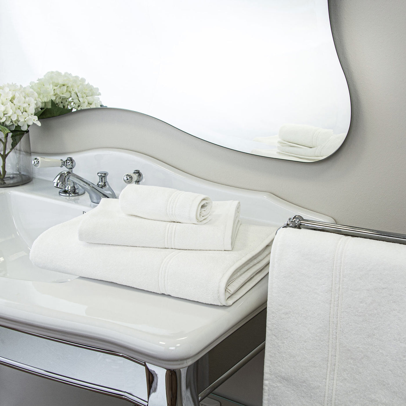 A Frette Classic Hand Towel from Frette hangs beside a bathroom sink topped with plush towels, a chrome faucet, a wash cloth, and a vase of white flowers near a large decorative mirror.