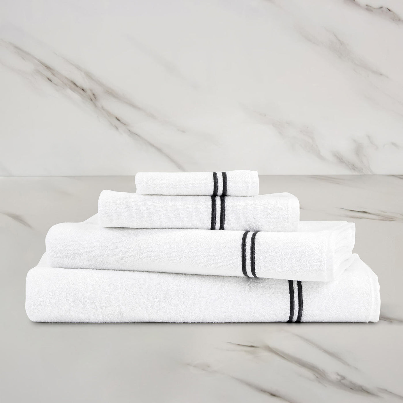 Four Frette Classic Hand Towels from Frette, neatly folded in white with black stripes, are stacked on a marble surface with a marble wall in the background.