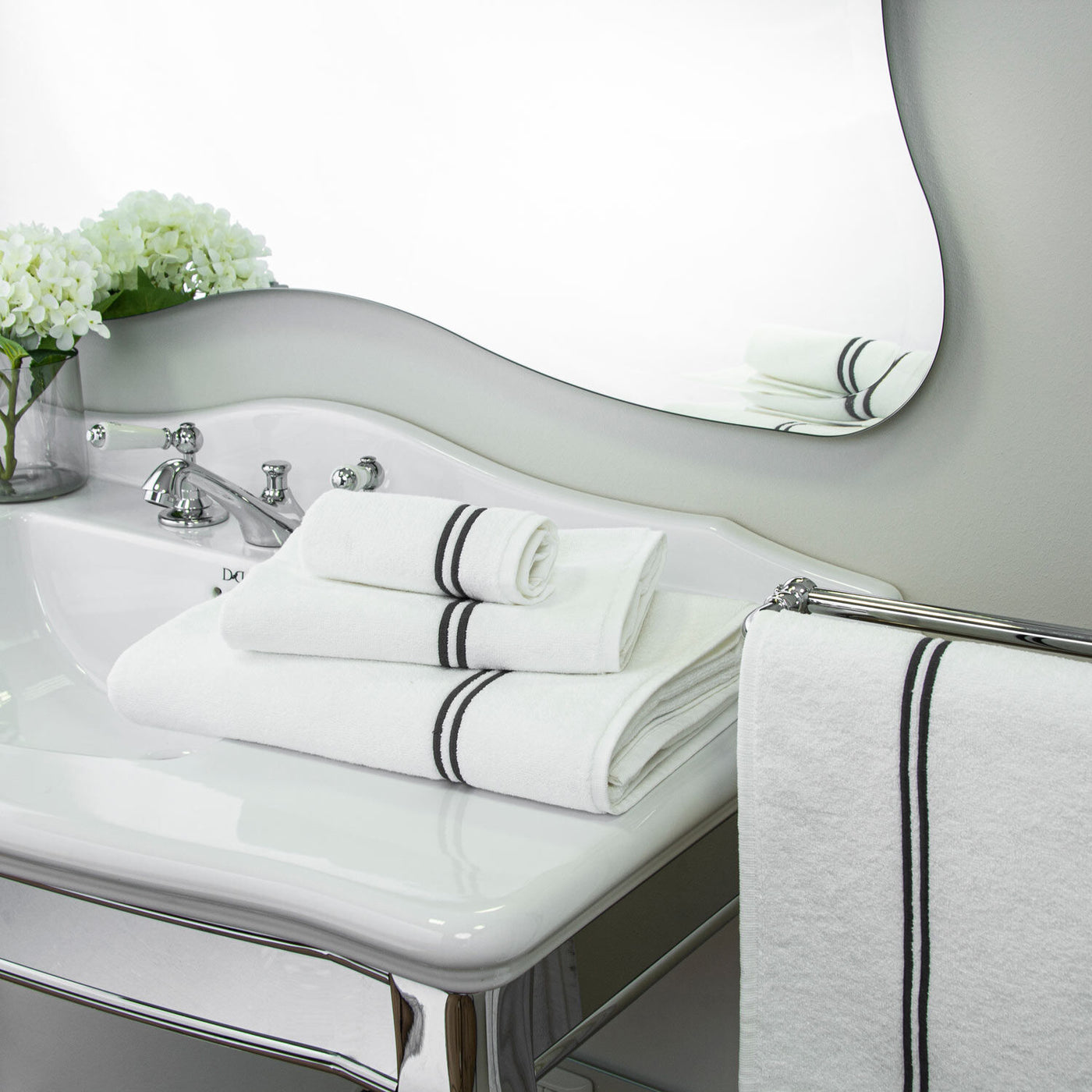 A white bathroom sink displays plush Frette Classic hand towels, neatly stacked with black stripes, a matching Frette towel on the rail, and a vase of white flowers by the faucet.
